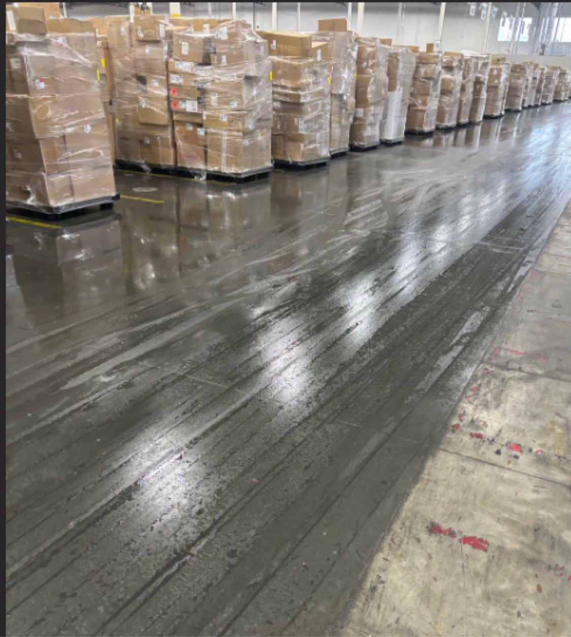 A dark treated traffic lane running beside untreated concrete in an occupied warehouse stacked with palletized freight