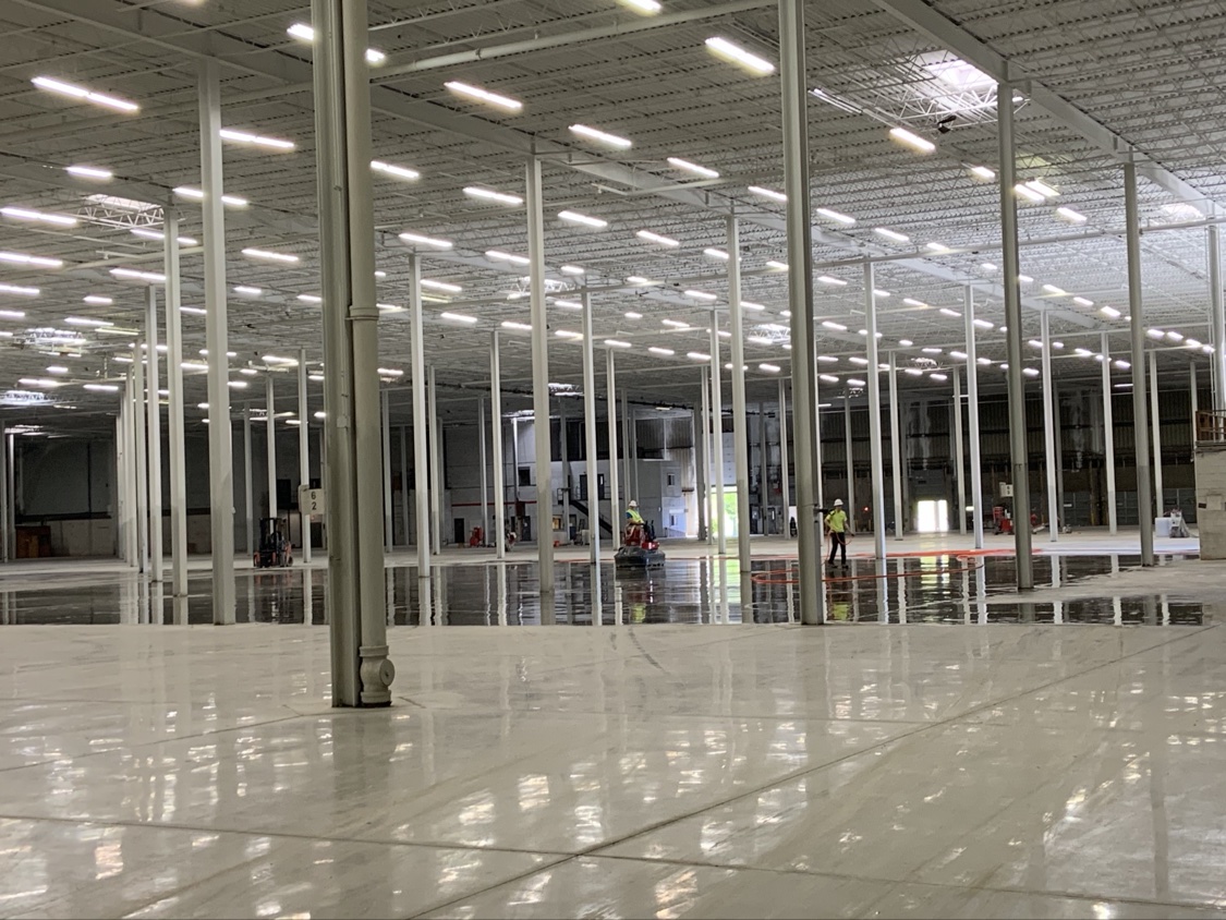 Vast white polished warehouse floor with SHMAK crew members applying finish in the distance between the columns